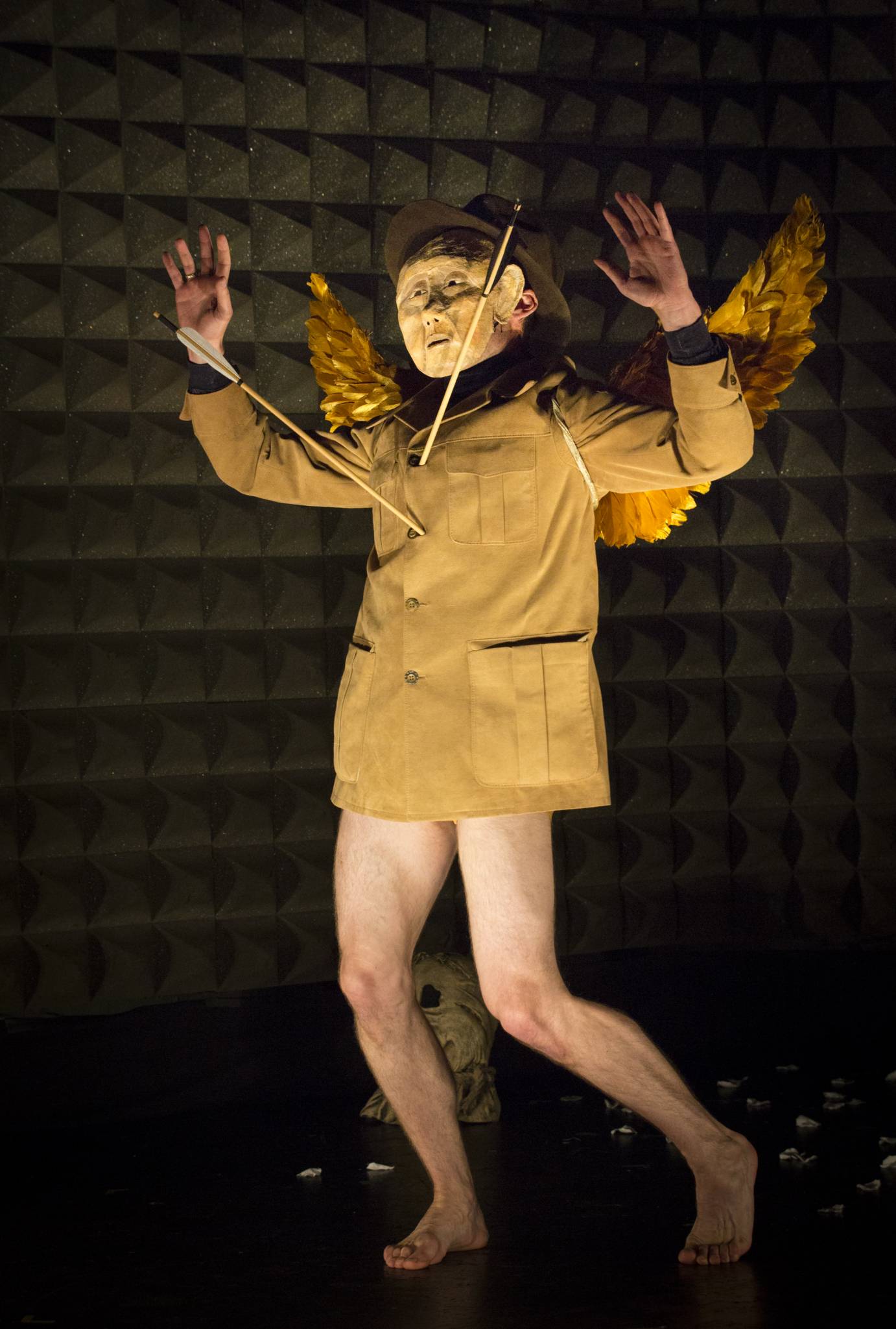 A man wears a mustard yellow jacket, no pants, his face obscured by a mask. 